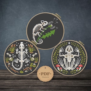 May include: Three cross-stitch embroidery hoops featuring skeletal designs. One depicts a lizard skeleton, another a chameleon skeleton, and the third a frog skeleton. The designs are on black fabric, with natural wood hoops and a PDF pattern tag.