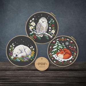 Könnte beinhalten: Drei runde Stickrahmen aus Holz mit Kreuzstichmotiven einer Schnee-Eule, eines Eisbären und eines Fuchses, jeweils umgeben von winterlichem Blattwerk. Ein kleines rundes Etikett mit dem Text PDF Pattern befindet sich zwischen den Rahmen.