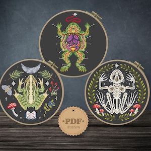 May include: Three cross-stitch embroidery hoops with dark gray backgrounds. One depicts a frog's anatomy, another a frog with a crescent moon, and the third a frog skeleton. A small tag with "PDF Pattern" is at the bottom.