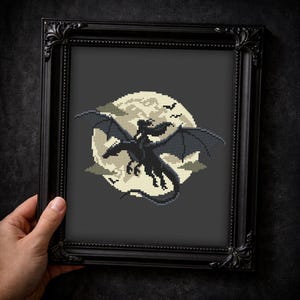 May include: A framed cross-stitch artwork featuring a black dragon silhouetted against a full moon. The dragon has outstretched wings, and the moon is surrounded by clouds and birds. The frame is black and ornate.