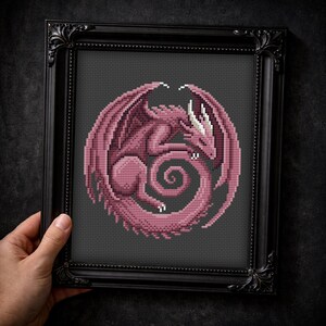 Peut inclure: Broderie au point de croix encadr&eacute;e repr&eacute;sentant un dragon rose dans un motif circulaire. Le dragon a des ailes et des cornes blanches, sur fond gris fonc&eacute;. Le cadre noir est orn&eacute; de d&eacute;tails.