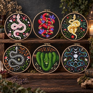 May include: Six circular embroidery hoops featuring cross-stitch designs on black fabric. The designs include a pink snake with flowers, a red and yellow snake with purple leaves, a yellow snake with foliage, a white snake skeleton, a green snake, and blue snakes with moon phases.