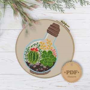 May include: Cross stitch pattern of a glass bottle with a cork stopper filled with a variety of succulents and small plants. The pattern is in a hoop and has a brown paper tag with the text "PDF Pattern".