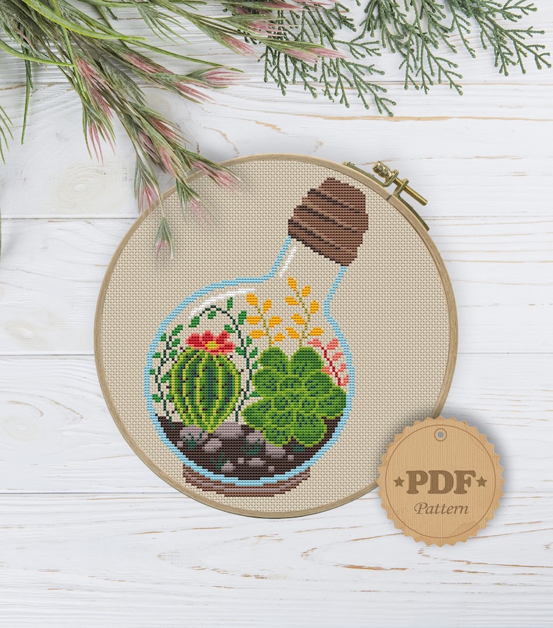 May include: Cross stitch pattern of a glass bottle with a cork stopper filled with a variety of succulents and small plants. The pattern is in a hoop and has a brown paper tag with the text "PDF Pattern".