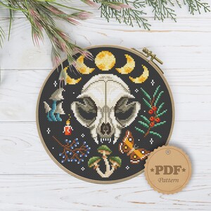 May include: A black and white cross stitch pattern featuring a skull with a crescent moon and stars. The skull is surrounded by mushrooms, berries, and a butterfly. The pattern is in a round wooden embroidery hoop. The text "PDF Pattern" is on a brown tag.