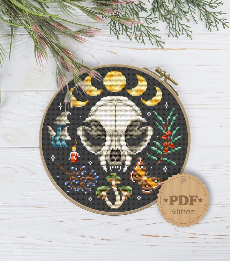 May include: A black and white cross stitch pattern featuring a skull with a crescent moon and stars. The skull is surrounded by mushrooms, berries, and a butterfly. The pattern is in a round wooden embroidery hoop. The text "PDF Pattern" is on a brown tag.
