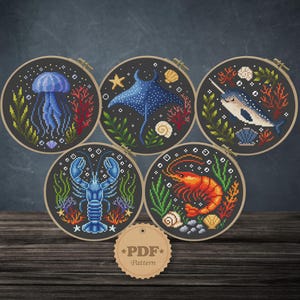 May include: Five cross-stitch patterns in wooden hoops, each featuring a different sea creature: a jellyfish, stingray, narwhal, lobster, and shrimp. The designs include coral, seaweed, and shells, all set against a dark background. A PDF pattern tag is at the bottom.