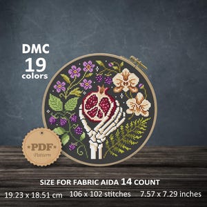 Puede incluir: Un patrón de punto de cruz en un bastidor de madera con una mano esquelética sosteniendo una granada partida, rodeada de flores moradas, moras y orquídeas blancas. Texto: DMC 19 colores, PDF Pattern, 19,23 x 18,51 cm.