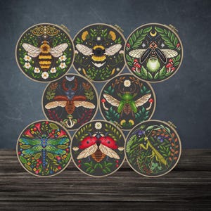 May include: Eight circular embroidery hoops, each displaying a different insect design. The collection includes a bee, bumblebee, firefly, beetle, and ladybug, all surrounded by detailed floral and leaf patterns. The colour scheme features greens, browns, and touches of red.