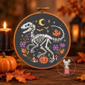 Può includere: Un ricamo a punto croce in un telaio di legno rotondo raffigurante uno scheletro di dinosauro bianco su sfondo grigio scuro. Il disegno include una mezzaluna gialla, pipistrelli arancioni, una foglia d'acero, fiori viola, funghi e una zucca di Halloween.