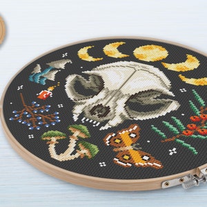 May include: A cross-stitch pattern featuring a white animal skull surrounded by moons, mushrooms, berries, and a butterfly. The pattern is on a black background and is framed in a wooden hoop.