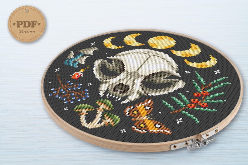 May include: A cross-stitch pattern featuring a white animal skull surrounded by moons, mushrooms, berries, and a butterfly. The pattern is on a black background and is framed in a wooden hoop.
