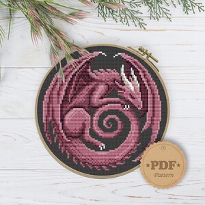 Peut inclure: Motif de point de croix d'un dragon rose aux ailes d&eacute;ploy&eacute;es, enroul&eacute; en cercle, sur fond gris fonc&eacute;. Le dragon est encadr&eacute; dans un cercle &agrave; broder en bois. Une &eacute;tiquette de motif PDF est attach&eacute;e.