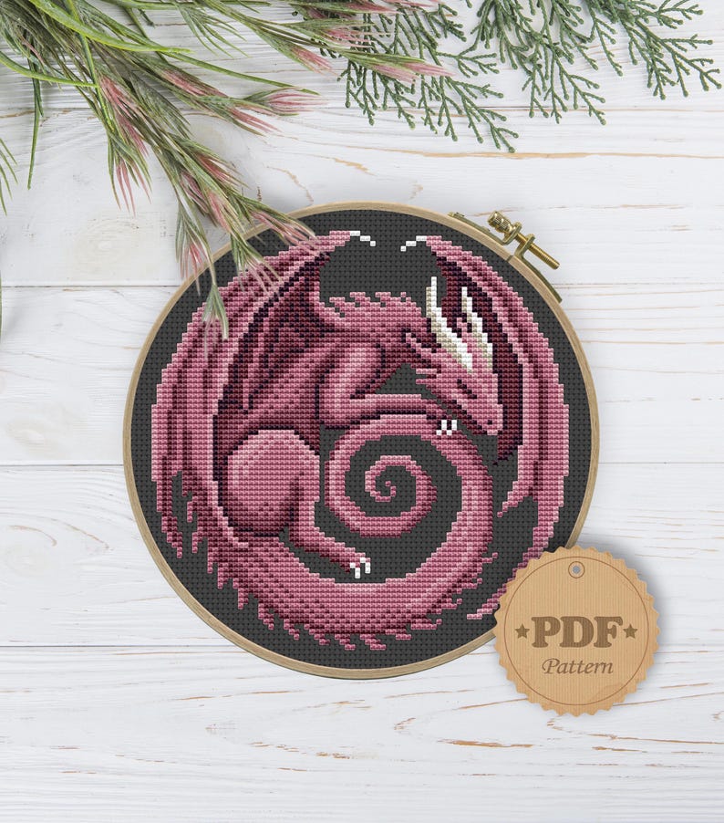 Peut inclure: Motif de point de croix d'un dragon rose aux ailes d&eacute;ploy&eacute;es, enroul&eacute; en cercle, sur fond gris fonc&eacute;. Le dragon est encadr&eacute; dans un cercle &agrave; broder en bois. Une &eacute;tiquette de motif PDF est attach&eacute;e.