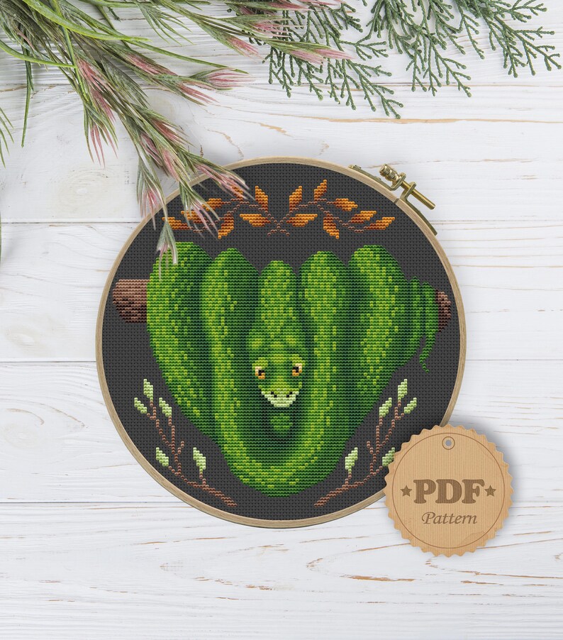 May include: A cross-stitch pattern featuring a green snake wrapped around a branch with leaves. The pattern is in a round embroidery hoop with a brown wooden frame. The text "PDF Pattern" is on a brown tag.