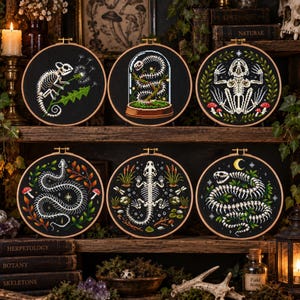 May include: Six circular embroidery hoops featuring white skeletal animal designs on black fabric. The designs include a chameleon with a dandelion, a snake in a glass dome, a frog with mushrooms, a coiled snake, a lizard, and a snake with a moon.