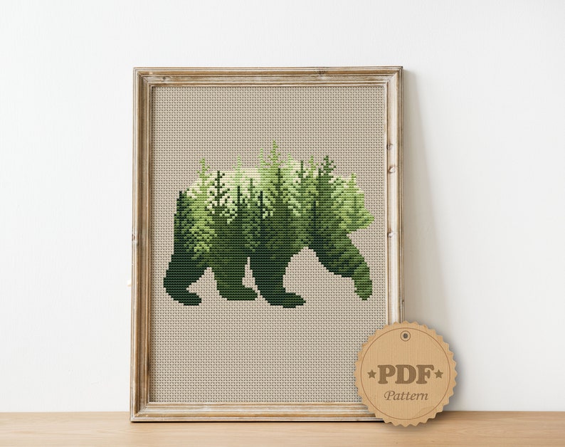 May include: Cross stitch pattern featuring a bear silhouette with a forest scene inside. The pattern is in a wooden frame and a brown tag with the text "PDF Pattern" is in the lower right corner.