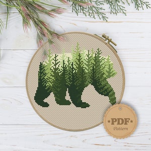 May include: A cross-stitch pattern of a bear silhouette with a forest scene inside. The pattern is in a round embroidery hoop and has a brown tag that says "PDF Pattern".