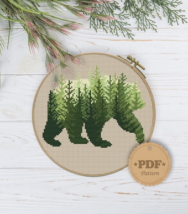 May include: A cross-stitch pattern of a bear silhouette with a forest scene inside. The pattern is in a round embroidery hoop and has a brown tag that says "PDF Pattern".