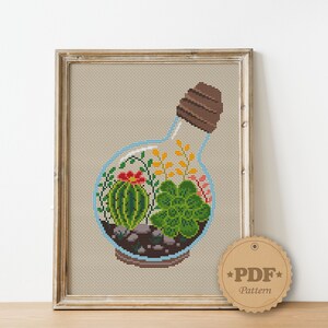 May include: Cross stitch pattern of a glass bottle terrarium with a cork stopper. The terrarium contains a variety of succulents, including a cactus, and small stones. The pattern is available as a PDF download.