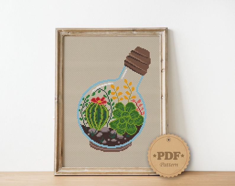 May include: Cross stitch pattern of a glass bottle terrarium with a cork stopper. The terrarium contains a variety of succulents, including a cactus, and small stones. The pattern is available as a PDF download.