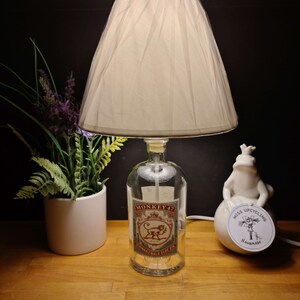Könnte beinhalten: Ein weißer Lampenschirm mit einer klaren Glasflaschenbasis. Die Flasche ist mit "Monkey 47 Schwarzwald Sloe Gin" beschriftet. Die Lampe steht auf einem Holztisch mit einer weißen Keramikfroschfigur und einem weißen Keramikpflanzentopf.