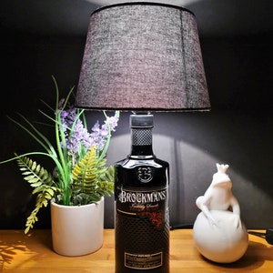 Könnte beinhalten: Ein schwarzer Lampenschirm mit einem schwarzen Stoffschirm sitzt auf einer durchsichtigen Glasflasche mit einem schwarzen Etikett, auf dem "Brockmans  Intensely Smooth Premium Gin" steht. Die Flasche steht auf einer Holzoberfläche mit einer weißen Keramikfroschfigur und einer Topfpflanze mit lila Blüten.