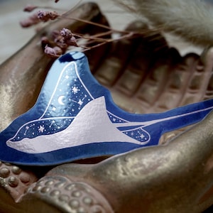 Può includere: Un adesivo blu e argento di una manta con stelle e una luna crescente. L'adesivo è su una superficie dorata.