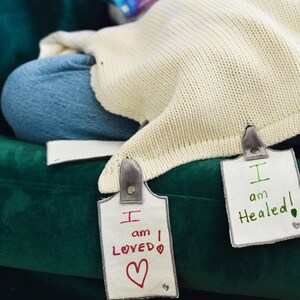 May include: Two white fabric tags with gray trim and black buttons. The tags have handwritten messages in red and green ink. The first tag says "I am LOVED" with a red heart. The second tag says "I am Healed!" with a green leaf.