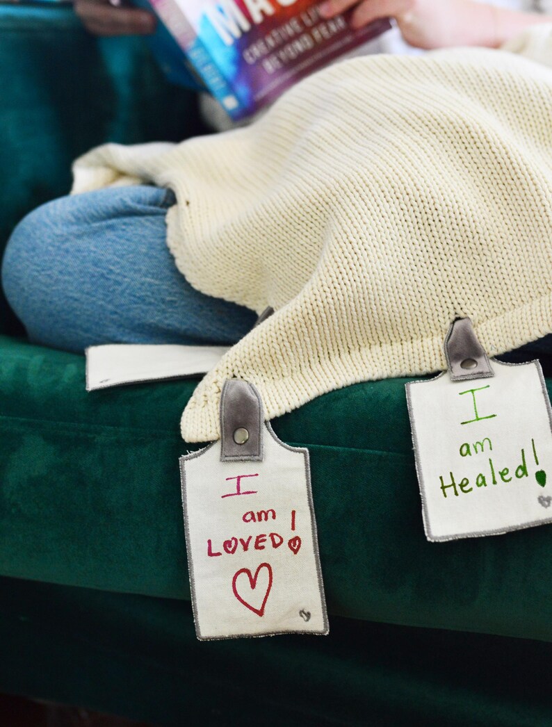 May include: Two white fabric tags with gray trim and black buttons. The tags have handwritten messages in red and green ink. The first tag says "I am LOVED" with a red heart. The second tag says "I am Healed!" with a green leaf.