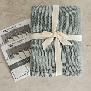 Puede incluir: Una manta de punto azul claro atada con una cinta crema. La manta se empaqueta con un juego de seis etiquetas personalizables que dicen "big hug" y "Personalizable Tags 6 Pieces".