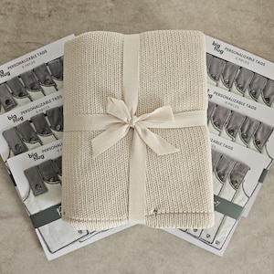 May include: A cream-colored knitted blanket with a white ribbon tied around it. The blanket is sitting on top of a stack of six personalized tags with the text "big hug" and "PERSONALIZABLE TAGS" printed on them.