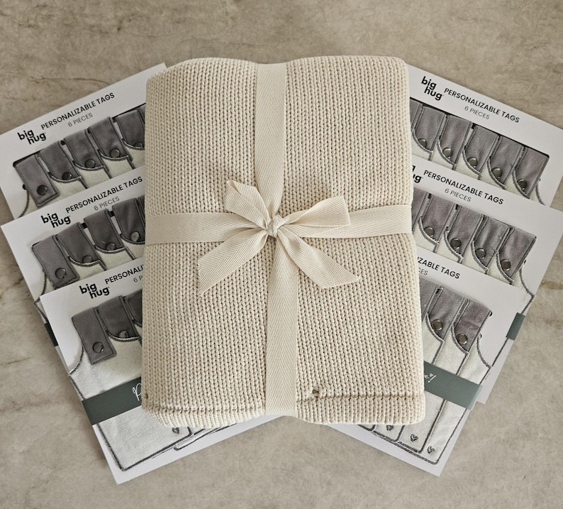 May include: A cream-colored knitted blanket with a white ribbon tied around it. The blanket is sitting on top of a stack of six personalized tags with the text "big hug" and "PERSONALIZABLE TAGS" printed on them.