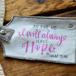 May include: A white fabric tag with gray stitching and a gray fabric loop. The tag has the text "As for me, I will always have hope. Psalm 71:14" written in black ink. The word "hope" is written in pink ink.