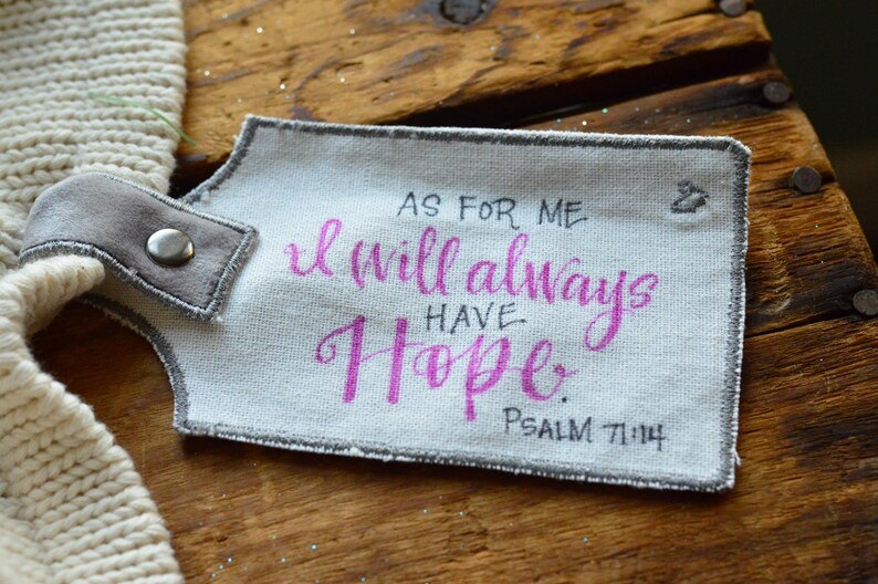 May include: A white fabric tag with gray stitching and a gray fabric loop. The tag has the text "As for me, I will always have hope. Psalm 71:14" written in black ink. The word "hope" is written in pink ink.