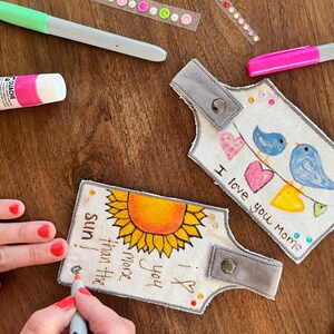 May include: Two fabric keychains with hand-painted designs. One keychain features a sunflower with the text "I love you more than the sun." The other keychain features two blue birds with hearts and the text "I love you Momie."