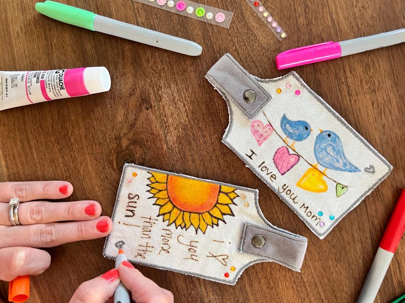 May include: Two fabric keychains with hand-painted designs. One keychain features a sunflower with the text "I love you more than the sun." The other keychain features two blue birds with hearts and the text "I love you Momie."