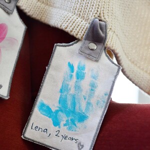 May include: A white fabric tag with a blue handprint and the text "Lena, 2 years". The tag is attached to a white knitted blanket with a gray fabric loop.