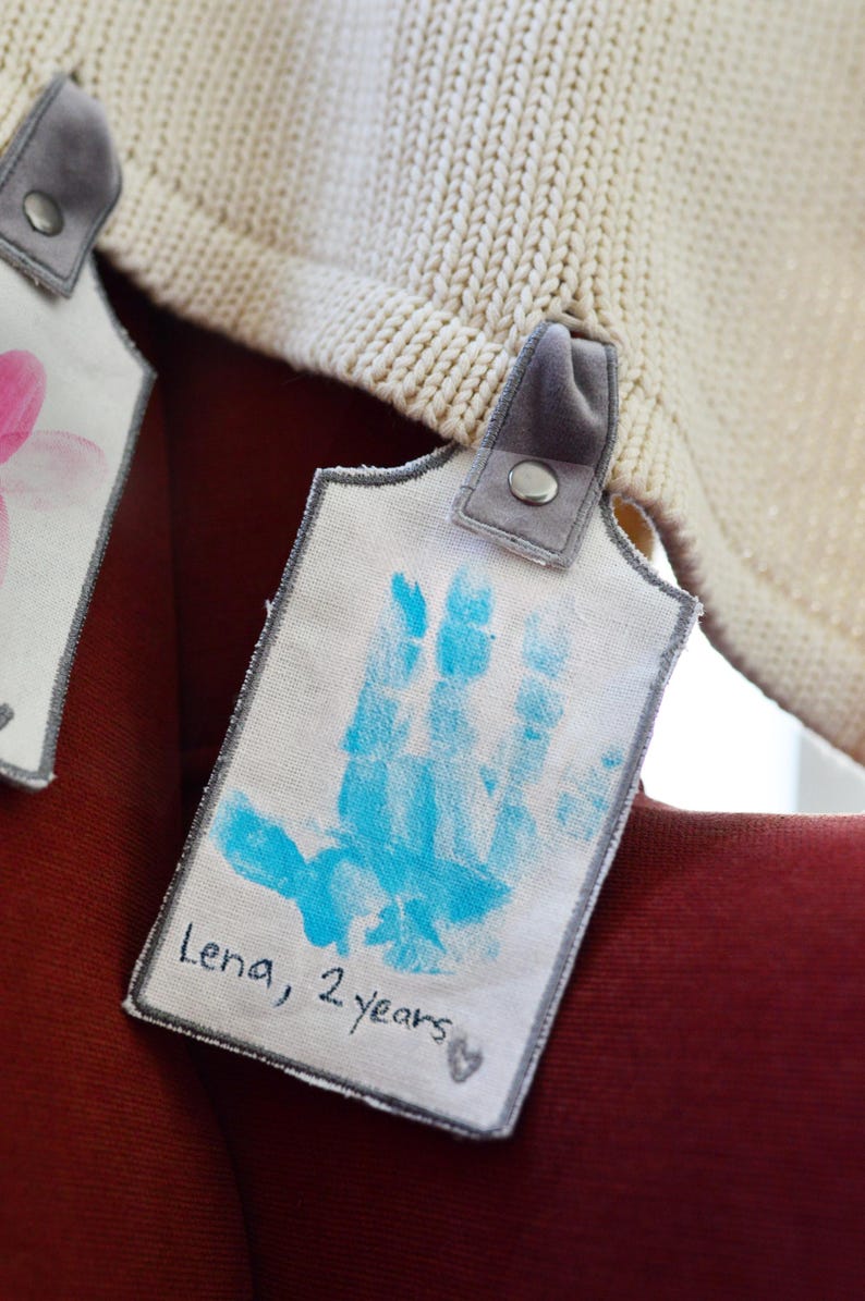 May include: A white fabric tag with a blue handprint and the text "Lena, 2 years". The tag is attached to a white knitted blanket with a gray fabric loop.