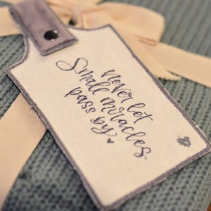 May include: A light blue knitted blanket with a white tag attached. The tag has a handwritten message that reads "Never let small miracles pass you by."