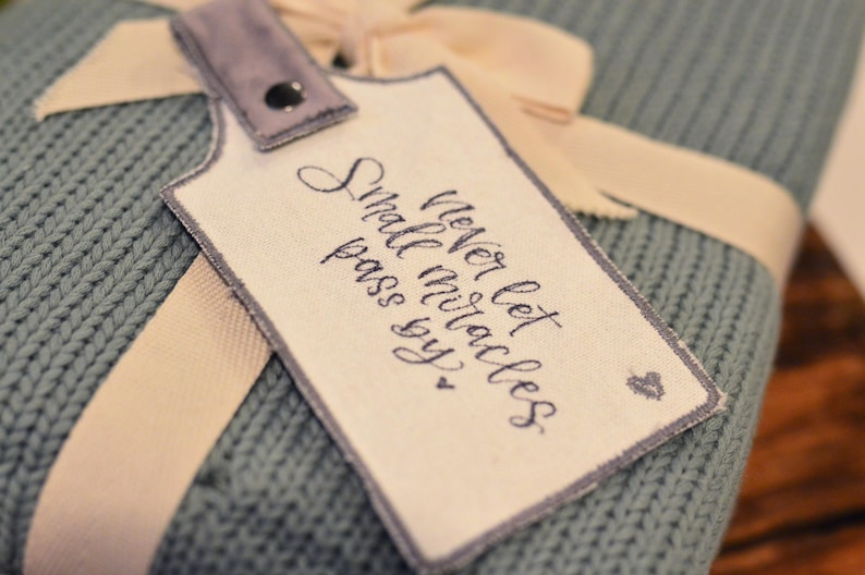 May include: A light blue knitted blanket with a white tag attached. The tag has a handwritten message that reads "Never let small miracles pass you by."