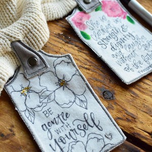 May include: Two fabric tags with inspirational quotes and floral designs. The first tag says "She is clothed in strength and dignity and laughs without fear of the future." The second tag says "Be gentle with yourself." Both tags have a gray loop for hanging.
