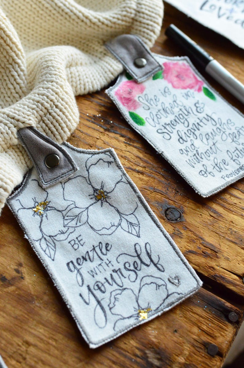 May include: Two fabric tags with inspirational quotes and floral designs. The first tag says "She is clothed in strength and dignity and laughs without fear of the future." The second tag says "Be gentle with yourself." Both tags have a gray loop for hanging.