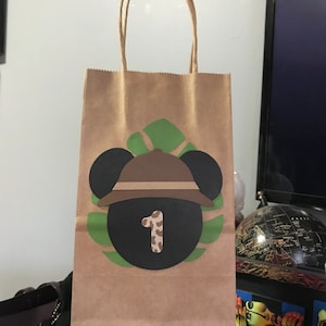 May include: A brown paper gift bag with a safari-themed design. The bag features a black circle with the number "1" in a cow print, a brown safari hat, and black mouse ears. Green leaf accents surround the design.