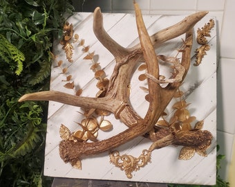 May include: A decorative wall hanging featuring deer antlers mounted on a white wooden plank. The antlers are a natural brown color, accented with gold-colored embellishments, including leaves and ornate scrollwork. The piece is a rustic, nature-inspired home decor item.