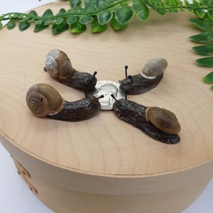May include: Four miniature snail figurines with brown and tan shells and dark bodies are arranged around a quarter coin on a light wood surface. The snails have detailed antennae and sit on a round wooden base.
