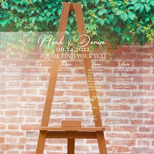 May include: A clear acrylic wedding seating plan with names and table numbers, presented on a wooden easel. The names "Noah & Denise" and the date "09.24.2022" are displayed. The background features a brick wall and green leaves.