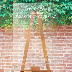 May include: Clear acrylic wedding seating plan with white calligraphy. The sign reads "Noah & Denise" and "Please Find Your Seat" with guest names listed under numbered sections. The sign is displayed on a wooden easel.