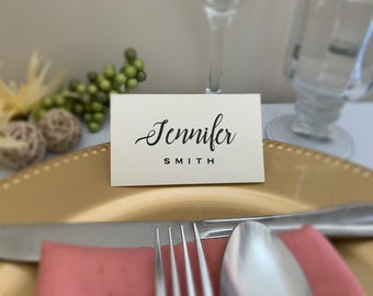 May include: A cream-colored place card with the name "Jennifer Smith" in elegant black script, set on a gold charger. The table setting includes silverware, a pink napkin, and decorative elements.