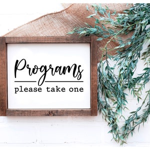 May include: A rectangular wooden sign with the words "Programs please take one" in black script. The sign is set against a white background, with a burlap cloth and green leafy garland in the background.